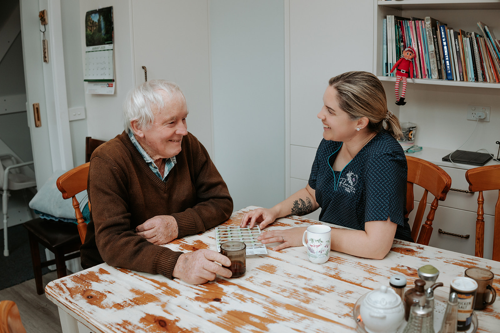 Aged care nurse supporting an elderly man with a medication check over a cup of tea at his kitchen table