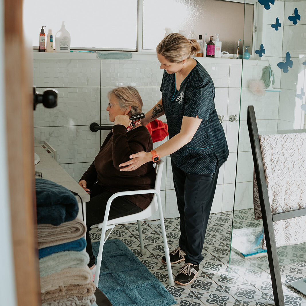 Nurse providing in-home support, helping an elderly woman with hair care in a bathroom.